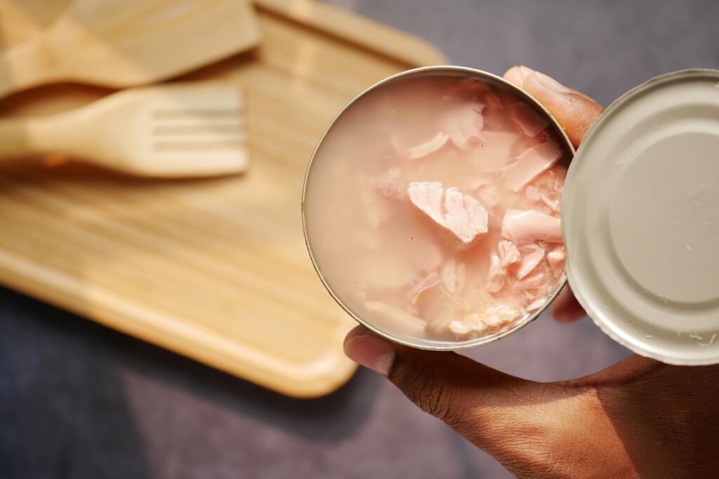 A close-up of an opened canned tuna held over a wooden cutting board and utensils.