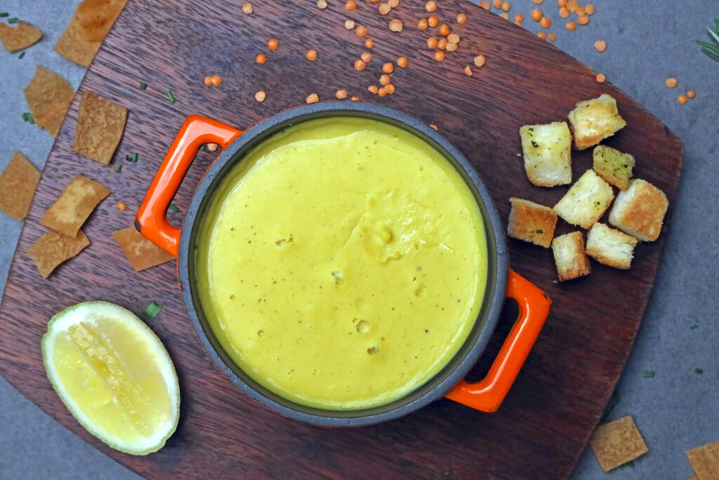 A top-down view of creamy yellow soup in an orange pot, garnished with croutons and served with lemon wedges.