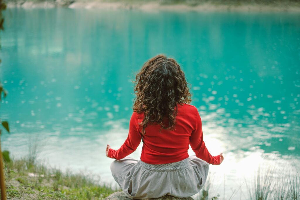 A woman in red meditates by a tranquil blue lake, reflecting calm and serenity.