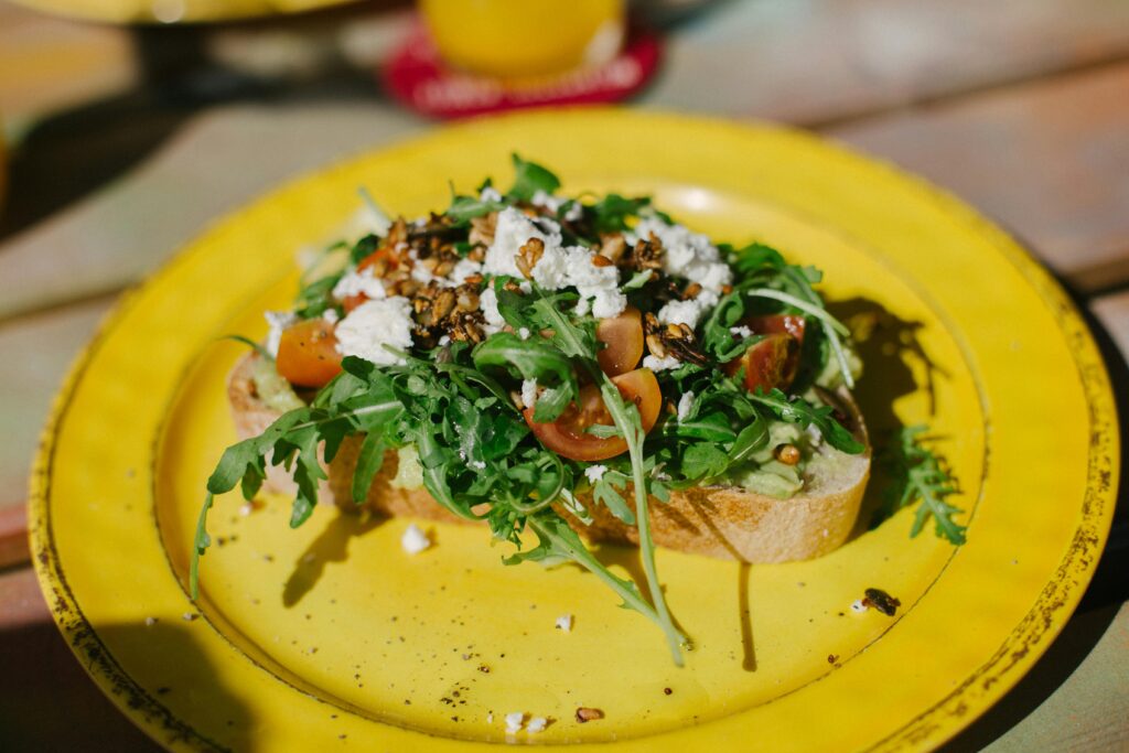 A vibrant and healthy avocado toast topped with cherry tomatoes, arugula, and feta cheese on a yellow plate.