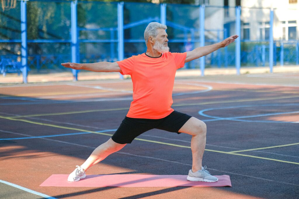 Elderly man performing warrior II yoga pose on an outdoor court.