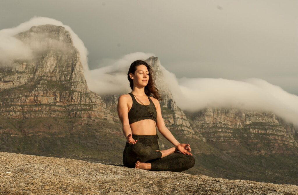 A woman practicing yoga and meditation outdoors in a scenic mountain setting.