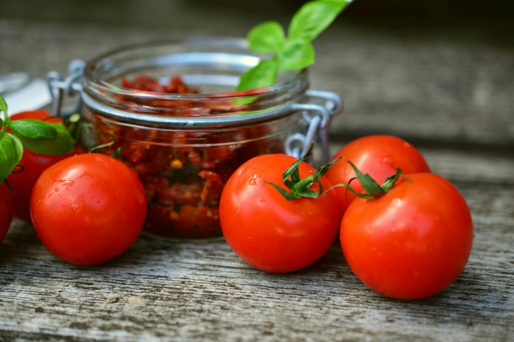 Close-up of fresh tomatoes beside a jar of sun-dried tomatoes on a rustic wooden surface.