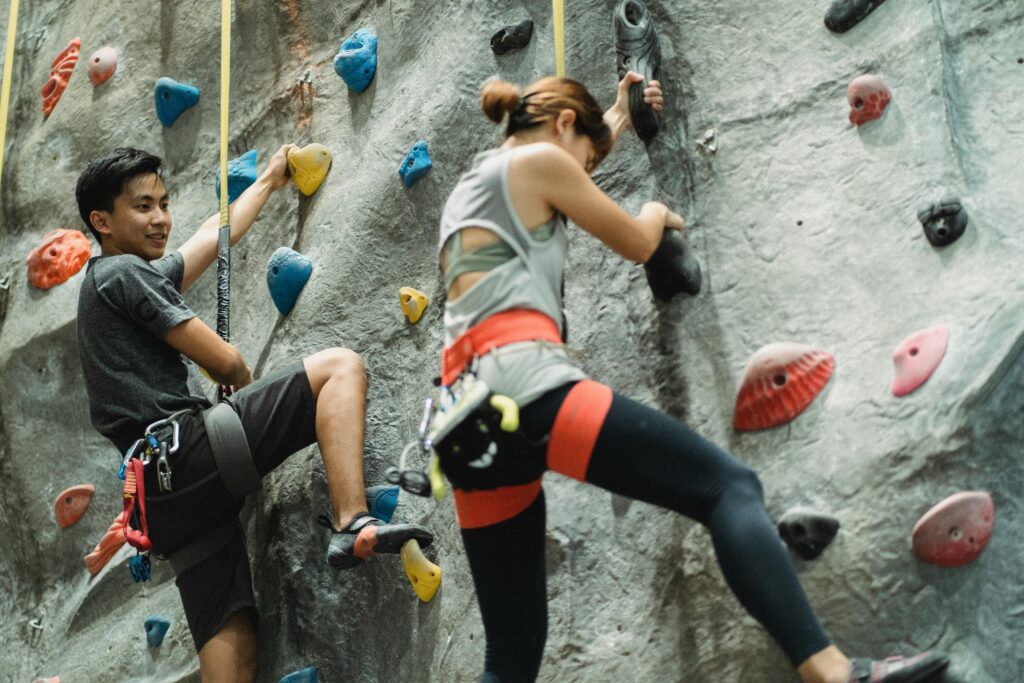 Low angle of young Asian male and female friends in sportswear and protective harness practicing bouldering together in climbing gym