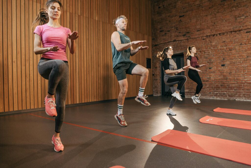 Four adults performing high knees in an indoor gym with wooden panel walls.