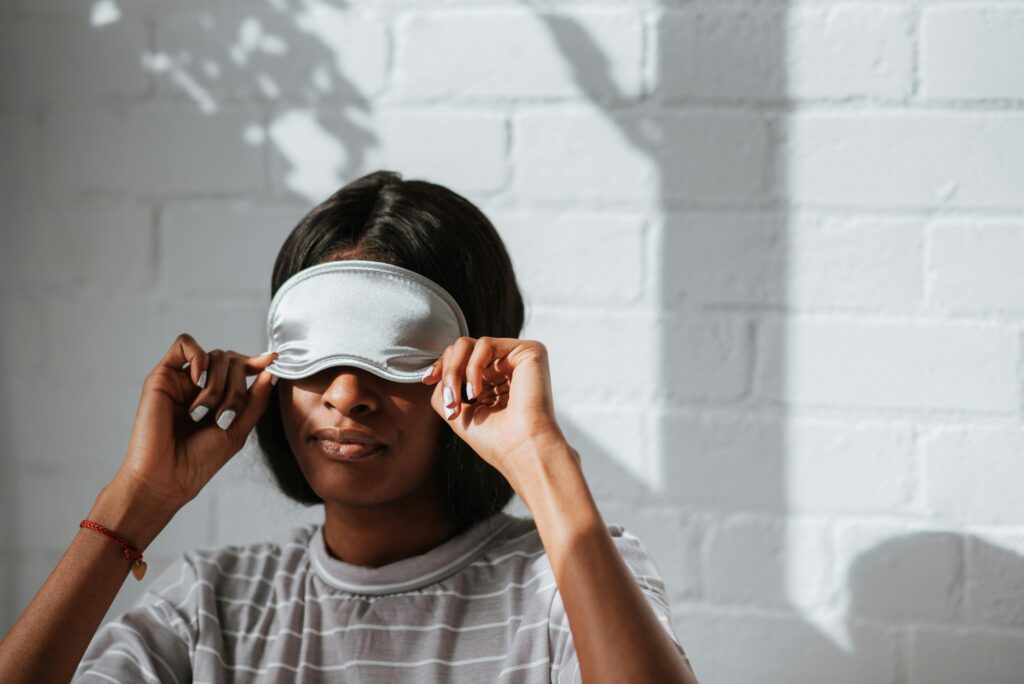 A woman wearing a grey striped shirt adjusts a sleeping mask in a bright indoor setting with shadows on the wall.